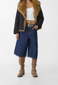 Zwarte pufferjas met bruine faux fur kraag, wit cropped topje, blauwe denim culottes en lichtbruine suède kniehoge laarzen met gespen.