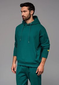 Felpa verde bluastro con cordini regolabili, maniche lunghe e tasca a marsupio. Tessuto testurizzato, con un piccolo logo giallo sulla manica.