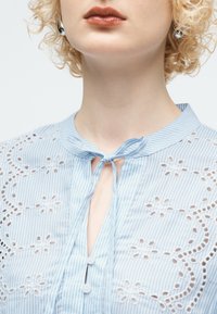 Κοντινή λήψη ενός ατόμου που φοράει μπλούζα με μπλε και λευκές ρίγες, διακοσμημένη με κεντήματα ματιών σε σχέδιο λουλουδιών και φιόγκο δεμένο στον λαιμό.