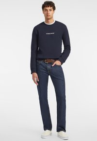 Tengerész színű pulóver, rajta "GUESS JEANS" logóval, sötétkék farmernadrággal és fehér sportcipővel párosítva. Kényelmes fazon, kerek nyakkivágással és hosszú ujjakkal.