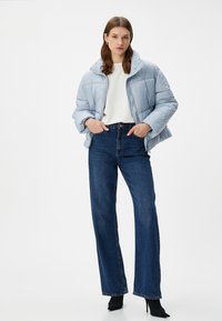 Chaqueta acolchada azul claro con cuello alto, cierre de cremallera y puños acanalados, combinada con jeans de tiro alto en azul oscuro y tacones negros.