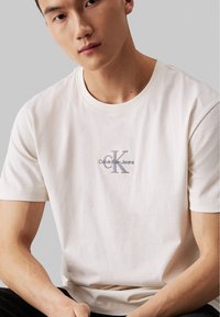 Baltas medvilninis marškinėliai su maža išsiuvinėta pilka logotipu, ant kurio užrašyta "Calvin Klein Jeans". Trumpi rankovės, apvalus kaklas ir įprastas kirpimas.