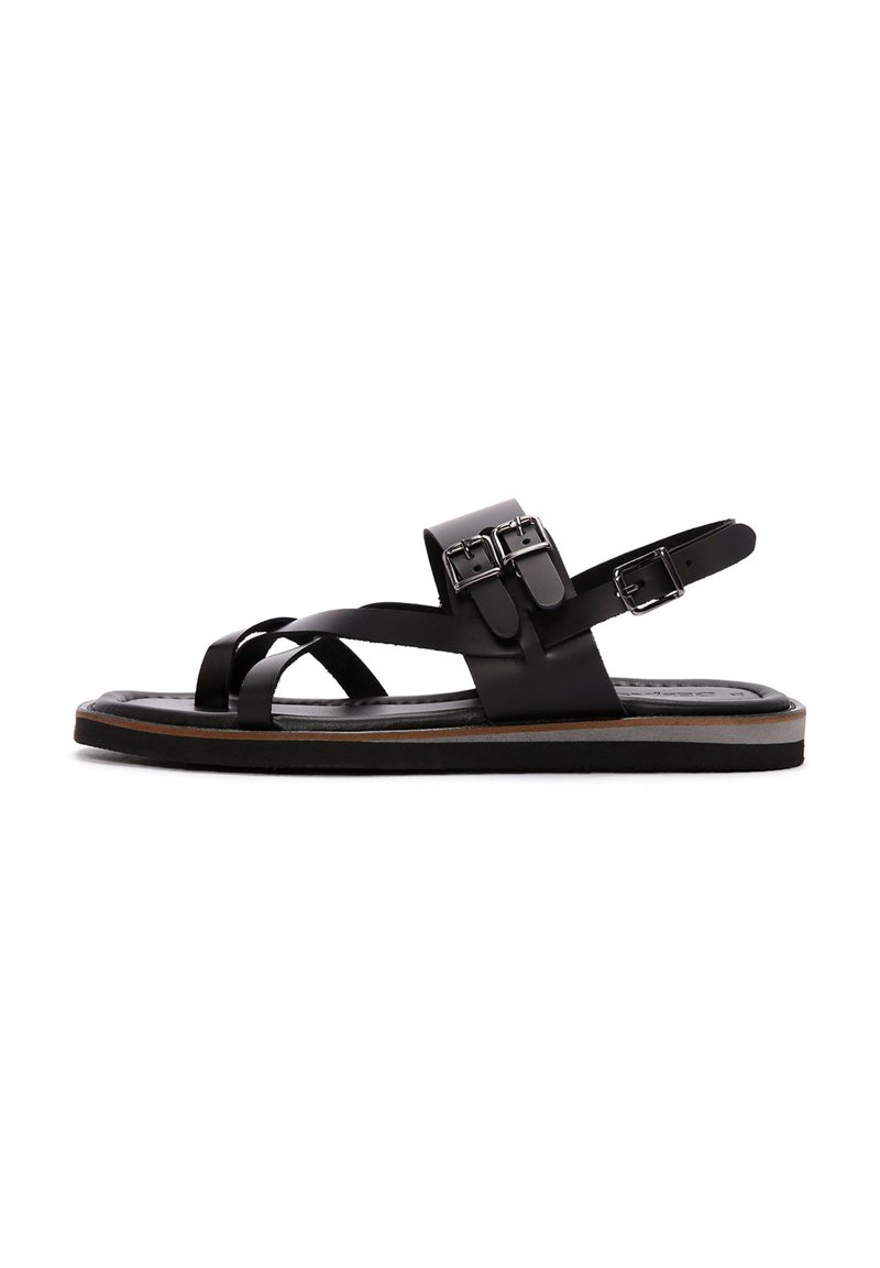 Las sandalias de cuero negro cuentan con correas entrecruzadas, cierre de hebilla ajustable y una suela plana con un ligero acento bronceado. Textura suave.