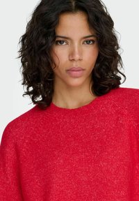 Rdeč pleten pulover z okroglim izrezom, raglanskimi rokavi in teksturirano površino. Tkanina je videti mehka in lahka.