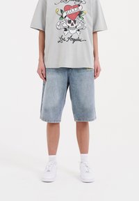 Camiseta de gran tamaño color gris claro con un gráfico de calavera colorido; pantalones cortos de mezclilla de longitud a la rodilla en azul con bordado amarillo en el dobladillo; zapatillas blancas.