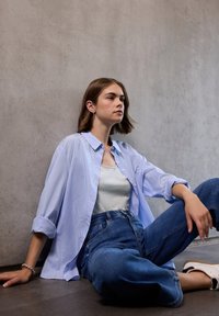 Camicia a righe azzurro chiaro con bottoni sopra una canotta grigia, abbinata a jeans blu a vita alta e sneakers bianche. Seduto su una superficie scura.