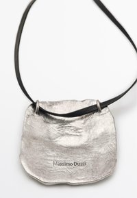 Pendentif métallique avec une forme texturée et irrégulière, attaché à un cordon en cuir noir. Présente la gravure "Massimo Dutti."