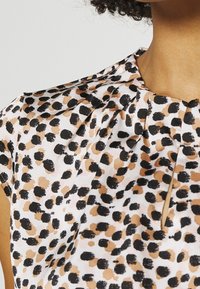 Kortärmad blus med vit bakgrund som har ett abstrakt mönster av svarta och bruna prickar, med plisserade detaljer vid halsringningen.
