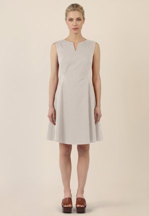 Frau mit blonden Haaren in einem knielangen, ärmellosen beigen Kleid und braunen Plateau-Sandalen mit offenem Zehenbereich, stehend vor einem einfarbigen beigen Hintergrund.