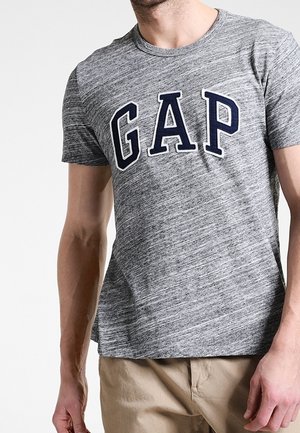 Mand iført en grå kortærmet GAP t-shirt og beige bukser, stående mod en ensfarvet lys baggrund.