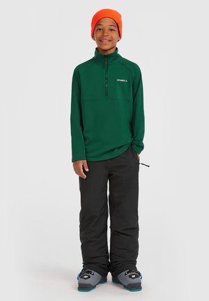 Grüne Fleecejacke mit Reißverschluss und O'Neill-Logo, kombiniert mit schwarzen Schneehosen und grauen Skischuhen. Das Model trägt eine orange Mütze.