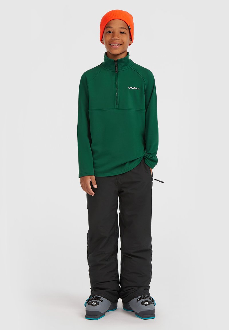 Groene fleece trui met rits en O'Neill-logo, gecombineerd met zwarte skibroek en grijze skischoenen. Model draagt een oranje muts.