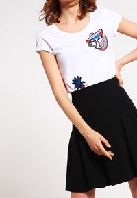 Valkoinen puuvillapaita, jossa on värikkäitä kirjontamalleja, yhdistetty mustaan laskostettuun hameeseen. Pehmeä kangas, pyöreä kaula-aukko, lyhyet hihat.