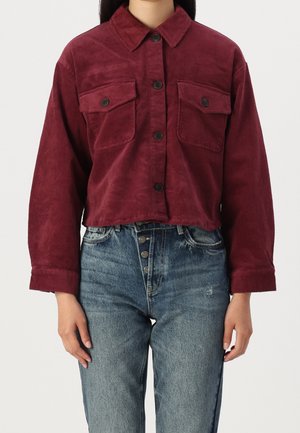 Veste en velours côtelé bordeaux avec un design court, dotée de deux poches sur la poitrine et de fermetures à boutons, associée à un jean bleu taille haute.