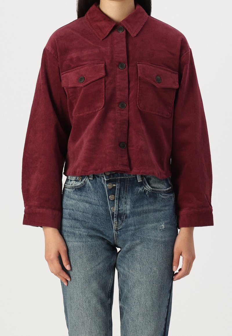 Bordeaux kordfløjlsjakke med en cropped design, der har to brystlommer og knaplukninger, parret med højtaljede blå jeans.