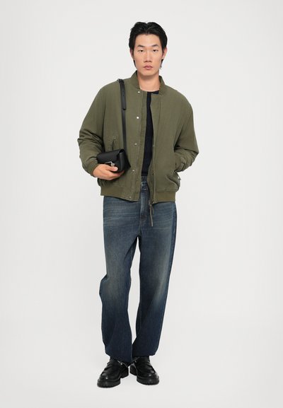 Grön bomberjacka, mörk tröja, lösa blå jeans och svarta skor. Håller en liten svart väska. Enfärgad vit bakgrund.