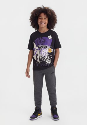 Enfant aux cheveux bouclés souriant, portant un T-shirt graphique de basket-ball noir des Los Angeles Lakers, un pantalon de survêtement gris Nike et des baskets noires, violettes et jaunes.