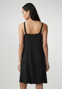Frau mit langen dunklen Haaren, die ein ärmelloses schwarzes Kleid trägt, steht mit dem Rücken zu einem schlichten hellgrauen Hintergrund.