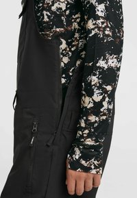 Zwarte overall met ritszakken en zijopeningen, gecombineerd met een lange mouwen top met een zwart, wit en bruin abstract patroon.