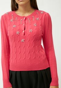 Roze kabelgebreide trui met een ronde halslijn, voorzien van decoratieve bloemversieringen en geribde manchetten enrand.