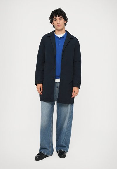Marinefrakke med knaplukning, båret over en blå sweater og hvid skjorte. Løst siddende blå jeans og sorte sko fuldender looket.
