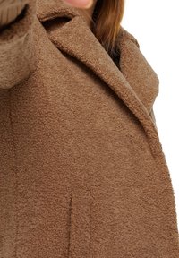 Brauner Fleece-Mantel mit strukturierter Oberfläche, breitem Revers und einreihigem Design. Sichtbare Nähte und keine Verschlüsse auf der Vorderseite.