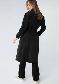 Trench coat in denim nero con vita regolabile, maniche lunghe e lunghezza fino al polpaccio, abbinato a pantaloni neri coordinati e scarpe da ginnastica bianche.