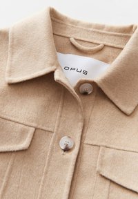 Beige wollen overhemdjasje met een kraag, twee voorzakken en ronde knopen. Etiket leest "OPUS." Zachte textuur is duidelijk zichtbaar in de stof.