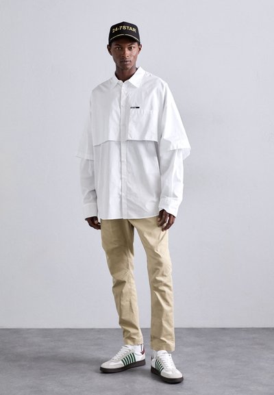 White oversized button-up shirt with a cropped front, beige trousers, and white sneakers with green stripes. Black cap with yellow text.