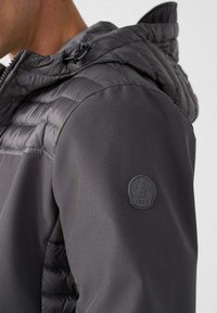 Grijze doorgestikte jas met een gladde textuur, afneembare capuchon en een rubberen logopatch op de linker schouder. Voorzien van een ritssluiting aan de voorkant.