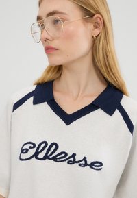 Młoda kobieta z długimi blond włosami, nosząca okulary w złotej oprawie oraz białą dzianinową bluzkę z granatowym kołnierzykiem i logo "Ellesse".