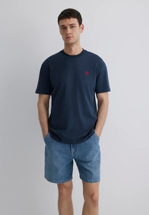 Himmelblaues Baumwoll-T-Shirt mit kurzen Ärmeln, rundem Halsausschnitt und einem roten Logo-Akzent auf der Brust, kombiniert mit Denim-Shorts.