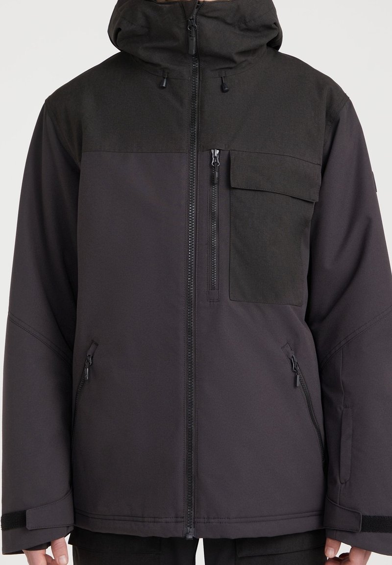 Veste à capuche noire et gris foncé avec fermeture éclair avant, poche poitrine avec rabat, deux poches latérales zippées et sangles ajustables aux poignets.