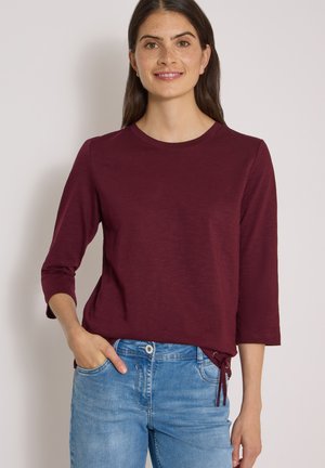 Femme aux longs cheveux foncés portant un haut bordeaux à manches trois quarts et un jean bleu, souriante avec une main dans la poche.