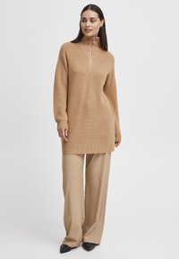 Robe pull en tricot beige avec un col montant et une fermeture éclair quart, associée à un pantalon large beige et des chaussures noires à bouts pointus.