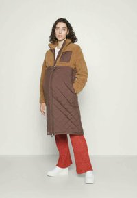 Καφέ επενδεδυμένο παλτό με μπεζ φλις λεπτομέρειες, ψηλό γιακά και κλείσιμο με φερμουάρ. Φοριέται πάνω από κόκκινες ριμπ παντελόνες και λευκά παπούτσια.