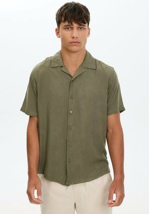 Joven con cabello corto y oscuro, vestido con una camisa verde oliva de manga corta abotonada y pantalones beige claro, de pie frente a un fondo liso.