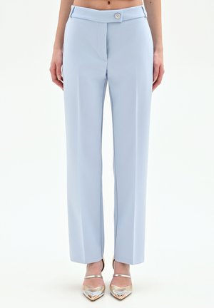 Donna con pantaloni su misura color azzurro chiaro a vita alta con bottoni frontali e tacchi argento a punta, in piedi contro uno sfondo neutro.