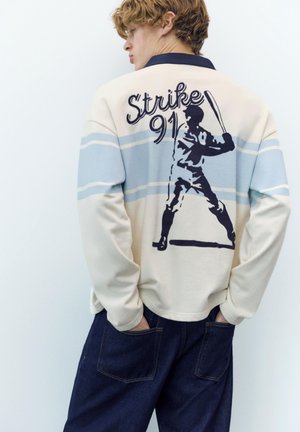Person trägt ein cremefarbenes und blau gestreiftes Langarmshirt mit der Aufschrift "Strike 91" und einer Grafik eines Baseballschlägers auf dem Rücken, die Hände in den Taschen einer dunklen Jeans.