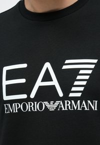 Μαύρο βαμβακερό μπλουζάκι με εμφανή λευκά λογότυπα "EA7" και "EMPORIO ARMANI", με ριγέ λεπτομέρεια στο "7". Απλός σχεδιασμός με στρογγυλή λαιμόκοψη.