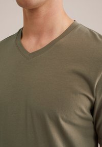 Olívazöld V nyakú póló, puha, sima anyagból készült, rövid ujjú és laza szabású. Finom, egyenletes textúrával rendelkezik.