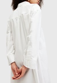 Persona in un vestito camicia bianco lungo a maniche lunghe con polsini abbottonati, in piedi con le mani incrociate dietro la schiena.