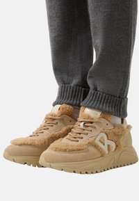 Beige Sneakers mit strukturierten Fellakzenten, Wildlederdetails und einer dicken Gummisohle. Kombiniert mit grauen, gerippten Jogginghosen mit Bündchen.