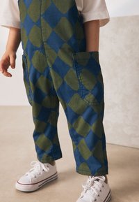 Salopette à losanges bleus et verts avec deux poches avant. Associée à des baskets blanches avec des accents rouges et des lacets rouges.