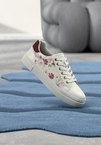 Baskets blanches avec motifs floraux roses, œillets dorés, et détail talon bordeaux flottant au-dessus d'un tapis bleu texturé.