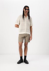Cremefarvet strikket kortærmet skjorte med åben meshmønster, parret med beige shorts og sorte sko, udstyret med runde solbriller og mellemlangt hår.