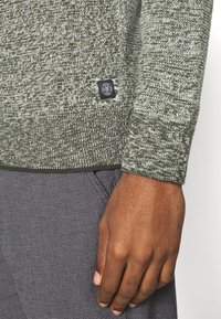Olivgrüner Pullover mit strukturiertem Strickmuster, mit einem schwarzen Label am Saum, getragen über karierten grauen Hosen.