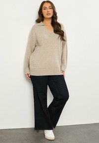 Beige gebreid sweater met een V-hals, geribbelde zoom en lange mouwen, gecombineerd met donkere jeans en witte sneakers. Zachte textuur, casual pasvorm.