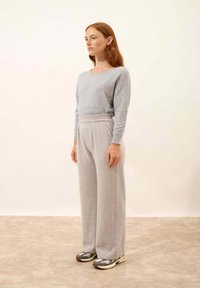 Světlý šedý kašmírový svetr s širokým výstřihem, v kombinaci s volnými světlými šedými kalhotami. Modelka má na sobě texturované tenisky.