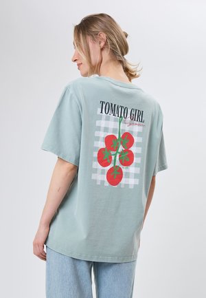 Femme portant un t-shirt surdimensionné bleu clair avec un motif de grappe de tomates rouges et le texte « TOMATO GIRL » dans le dos, associé à un jean bleu clair.